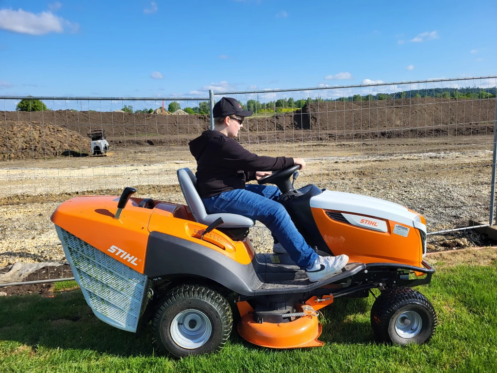 Orange-wei&szlig;er STIHL Aufsitzm&auml;her auf Wiese, Person in schwarzer Kleidung m&auml;ht Rasenfl&auml;che neben Baustellen-Zaun