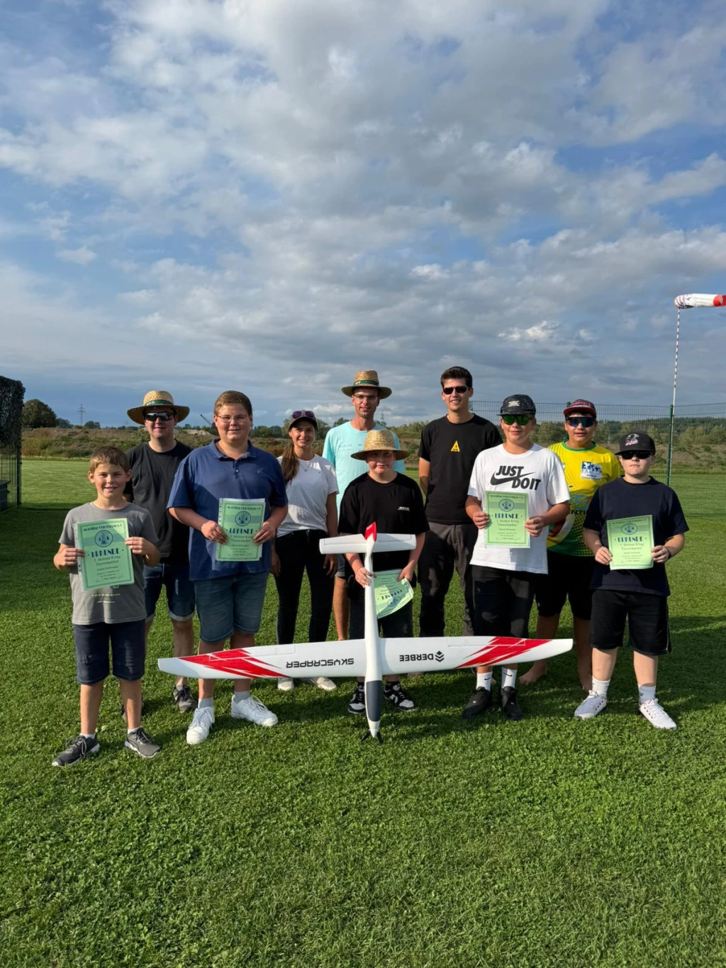 Elf Personen unterschiedlichen Alters mit Modellflugzeug auf grünem Rasenplatz, bewölkter Himmel, Urkunden in Händen