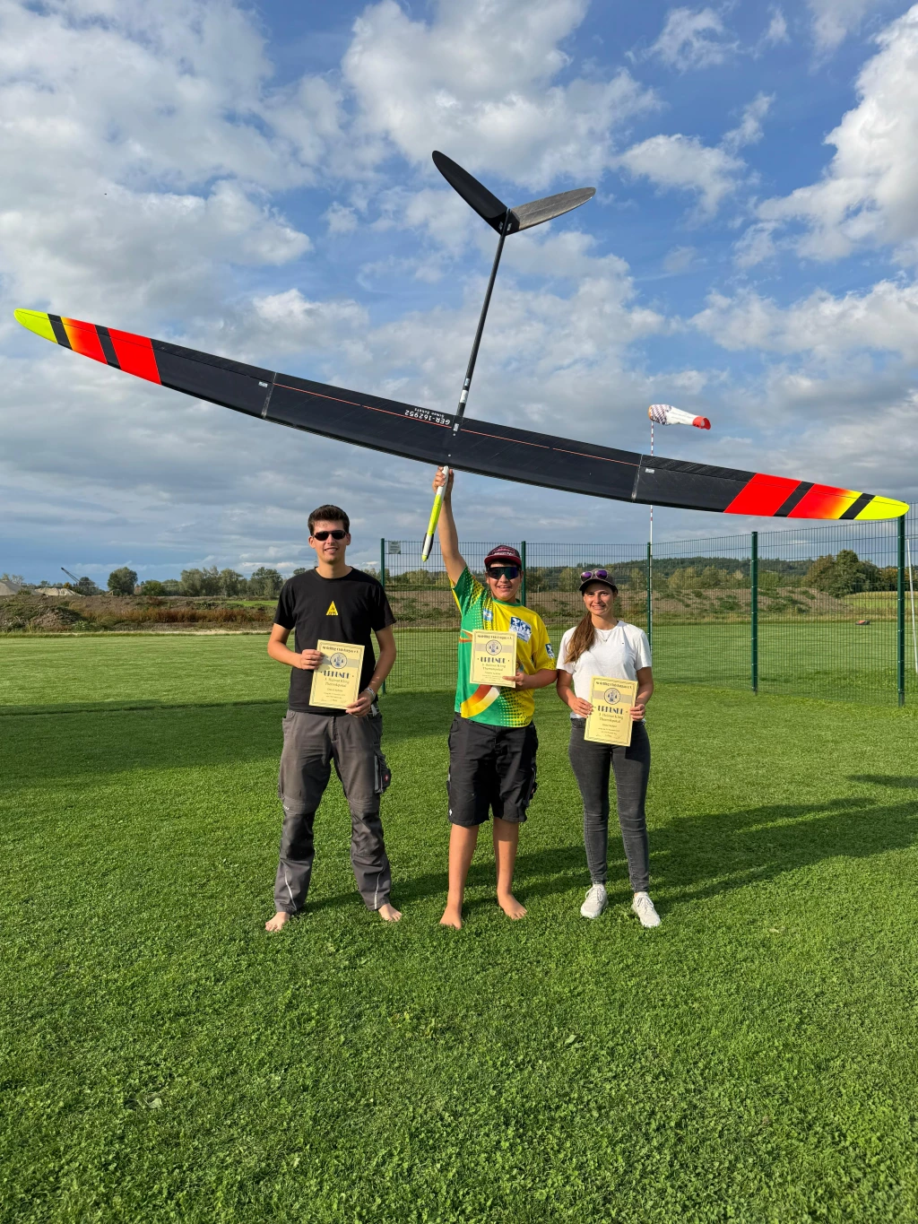 Drei Modellflieger mit Segelflugmodell, Auszeichnungen und Urkunden auf Vereinsgelände bei sonnigem Wetter