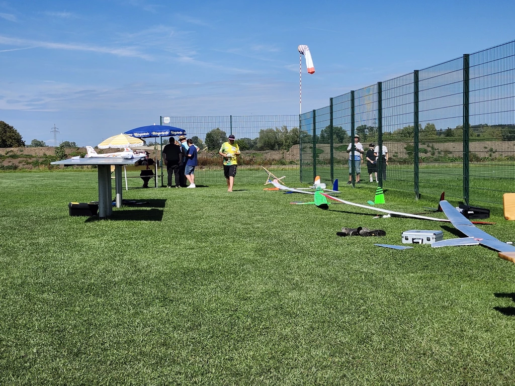 Grüne Rasenfläche mit mehreren Segelflugmodellen, Personen und Windfahne bei Modellflug-Event