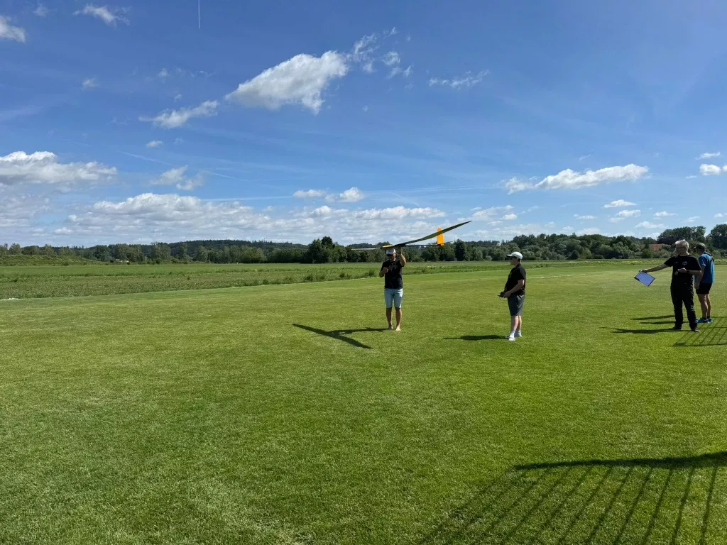 Segelflugmodell steigt nach dem Handstart über der Flugwiese, Pilot und Helfer schauen ihm nach