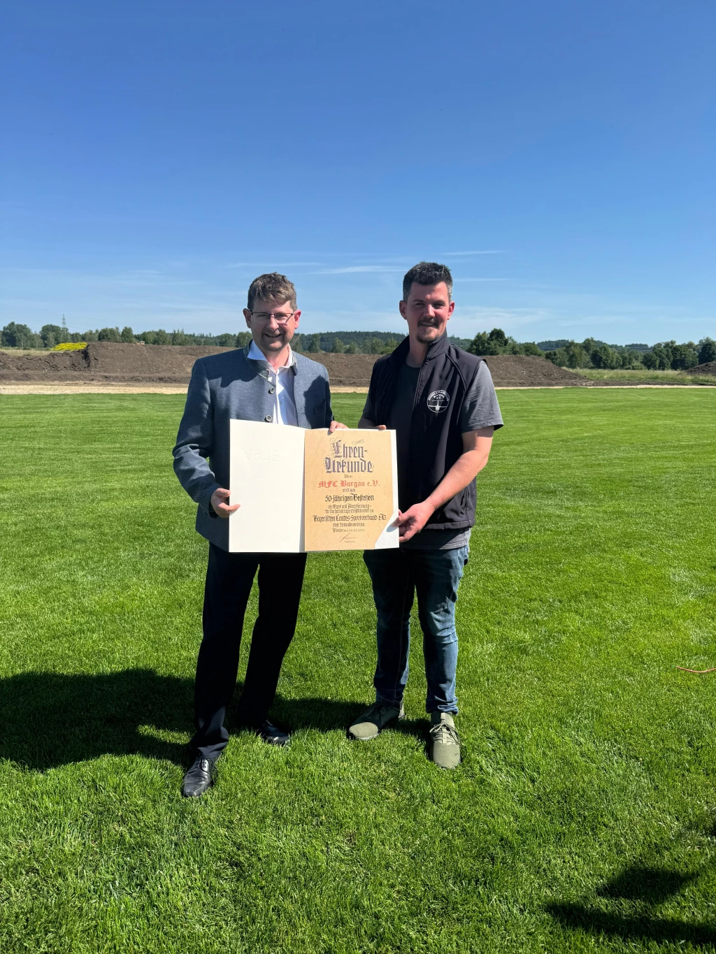 Zwei Männer in Vereinskleidung stehen auf grünem Rasenplatz und halten eine gelbe Ehrenurkunde
