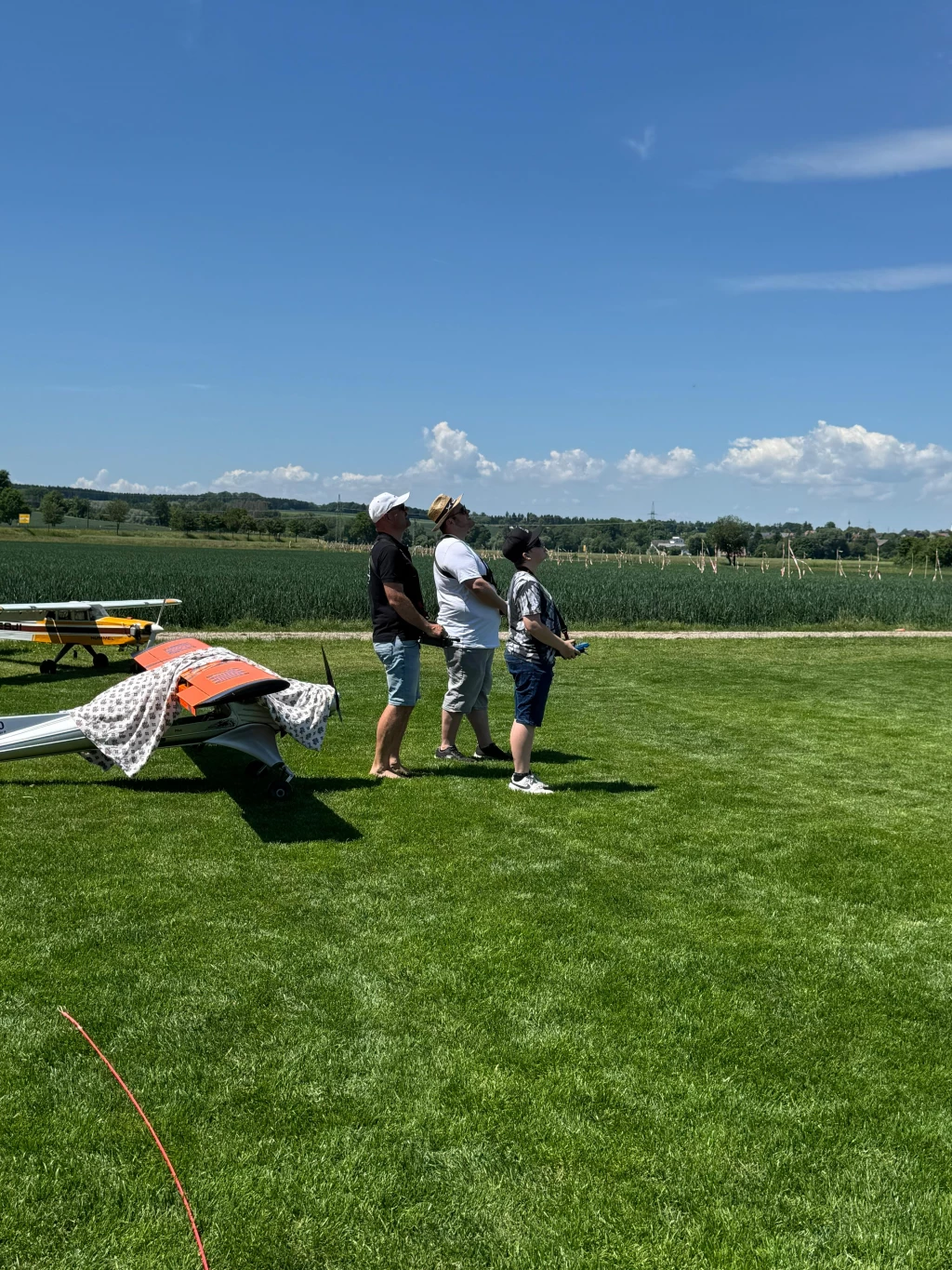 Drei Modellflug-Enthusiasten stehen auf grüner Rasenfläche neben Modellflugzeugen, Landschaft im Hintergrund