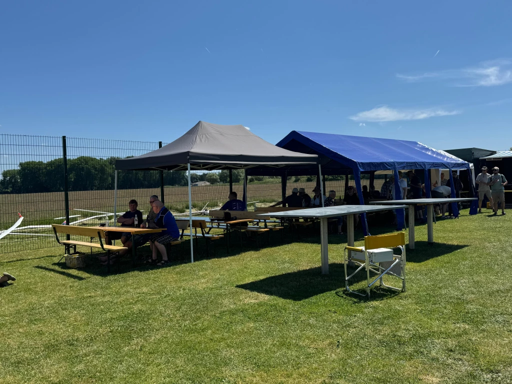 Zwei Pavillons auf Wiese, Tische und Stühle unter grauem und blauem Zelt, Zaun im Hintergrund