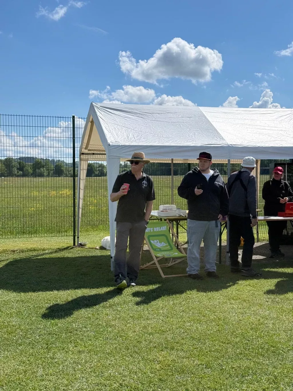 Teilnehmer stehen mit Getränken am Pavillon, davor ein Liegestuhl im Gras