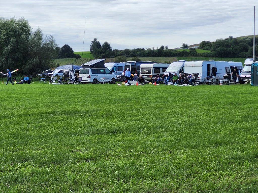 Mehrere Wohnwagen und Campingfahrzeuge parken auf einer grünen Wiese mit Bäumen im Hintergrund
