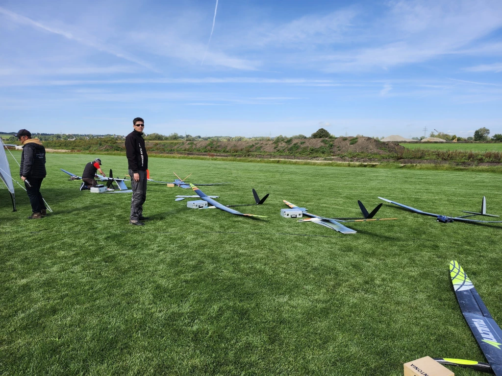 Mehrere Segelflugmodelle auf gr&uuml;ner Rasenfl&auml;che, drei Personen im Hintergrund, blauer Himmel mit Kondensstreifen