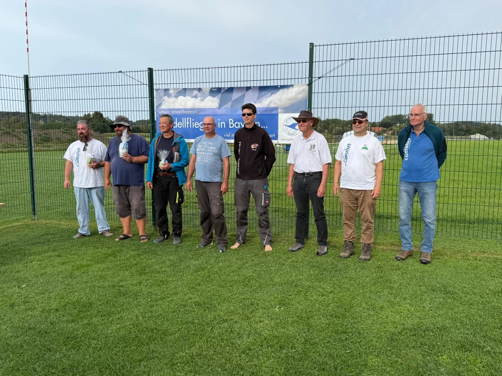 Acht Modellflug-Teilnehmer stehen in Reihe auf grünem Rasenplatz vor blauem Banner beim F5J-Wettbewerb