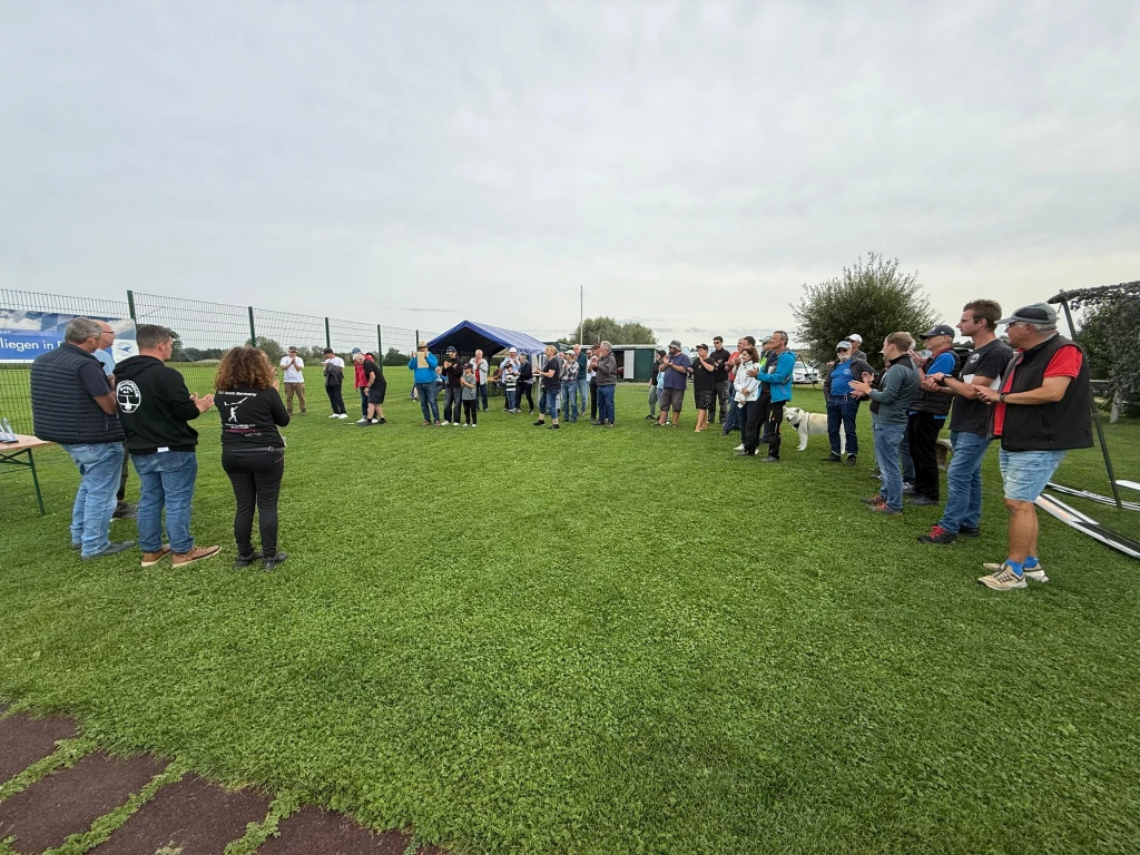 Gro&szlig;e Gruppe von Menschen in Freizeitkleidung steht in einem Halbkreis auf gr&uuml;nem Rasenplatz, blaues Zelt im Hintergrund