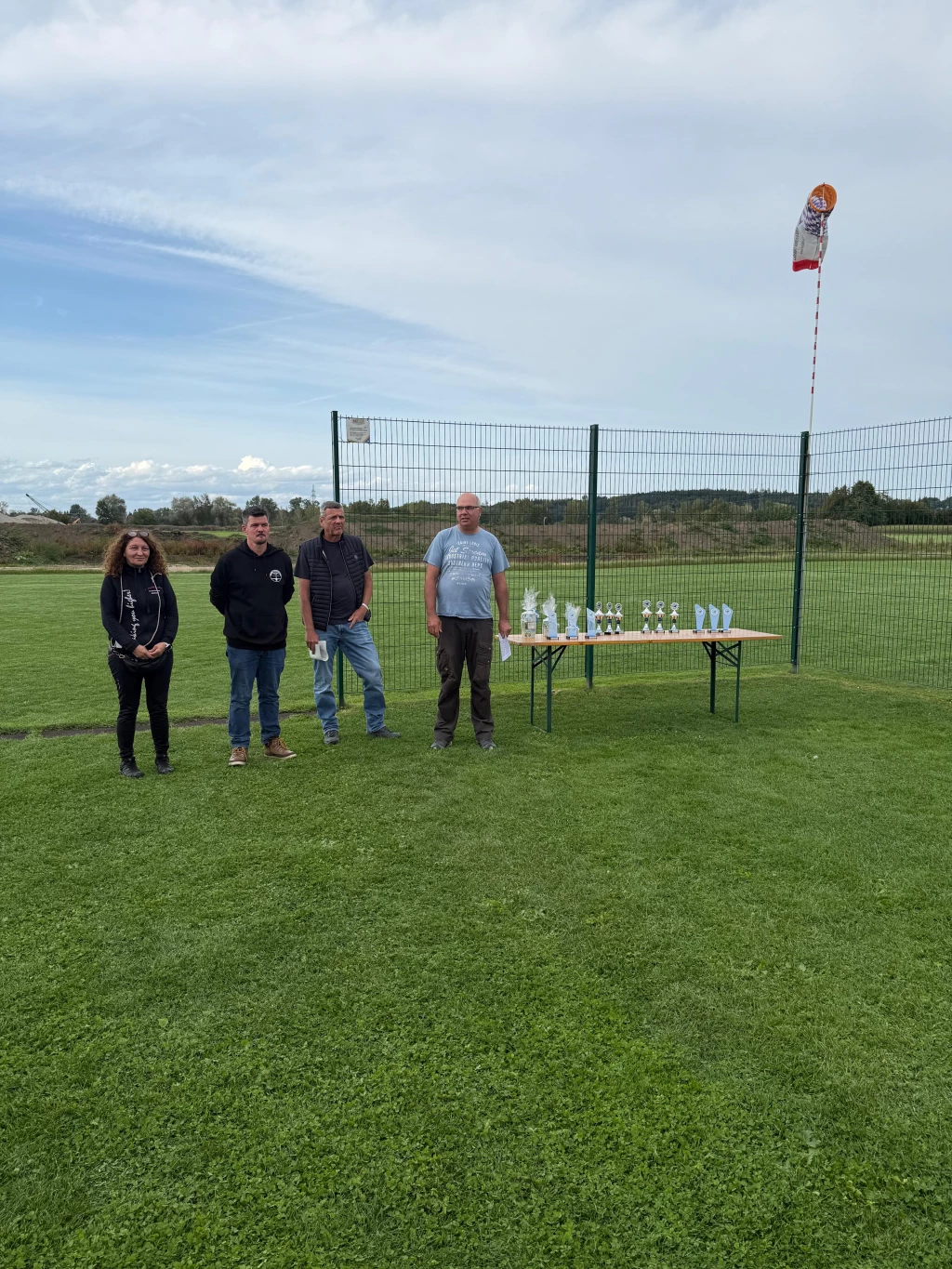 Vier Personen stehen auf grünem Rasenplatz neben Tisch mit Pokalen, Windfahne im Hintergrund