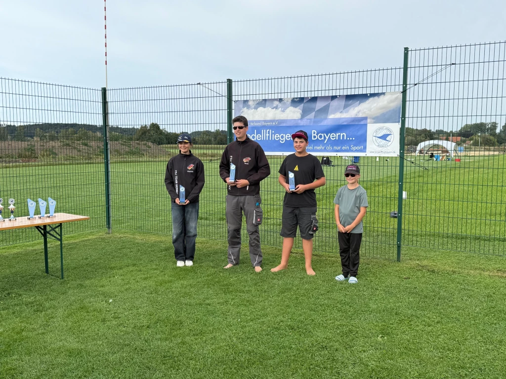Vier Personen bei Modellflug-Wettbewerb stehen auf Rasenplatz vor blauem Banner, drei mit Pokalen