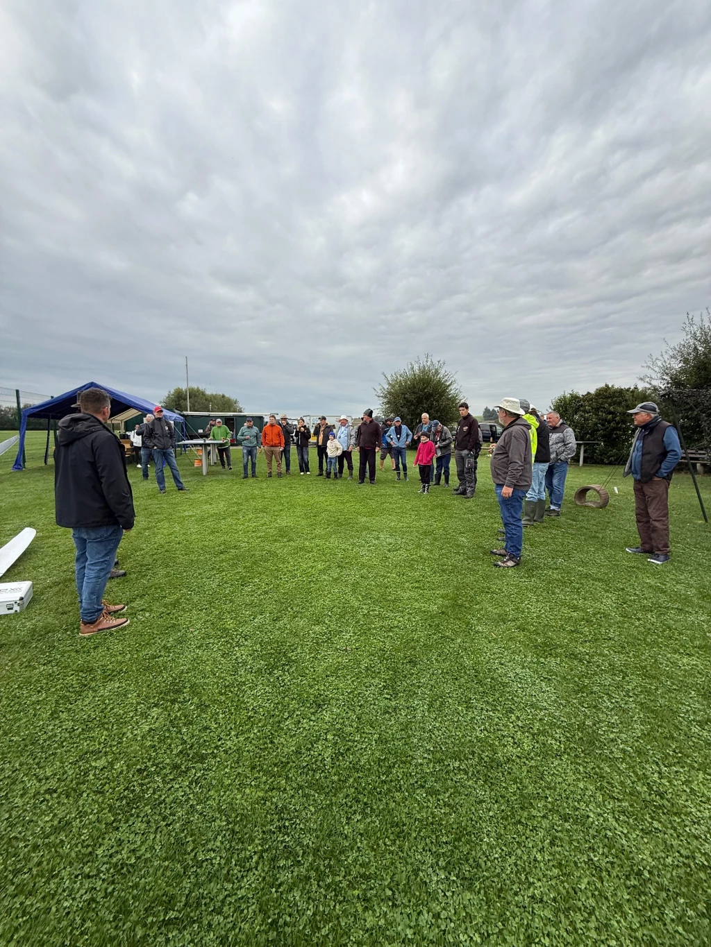 Gruppe von Personen auf grüner Wiese bei Modellflug-Veranstaltung, blaues Zelt im Hintergrund, bewölkter Himmel