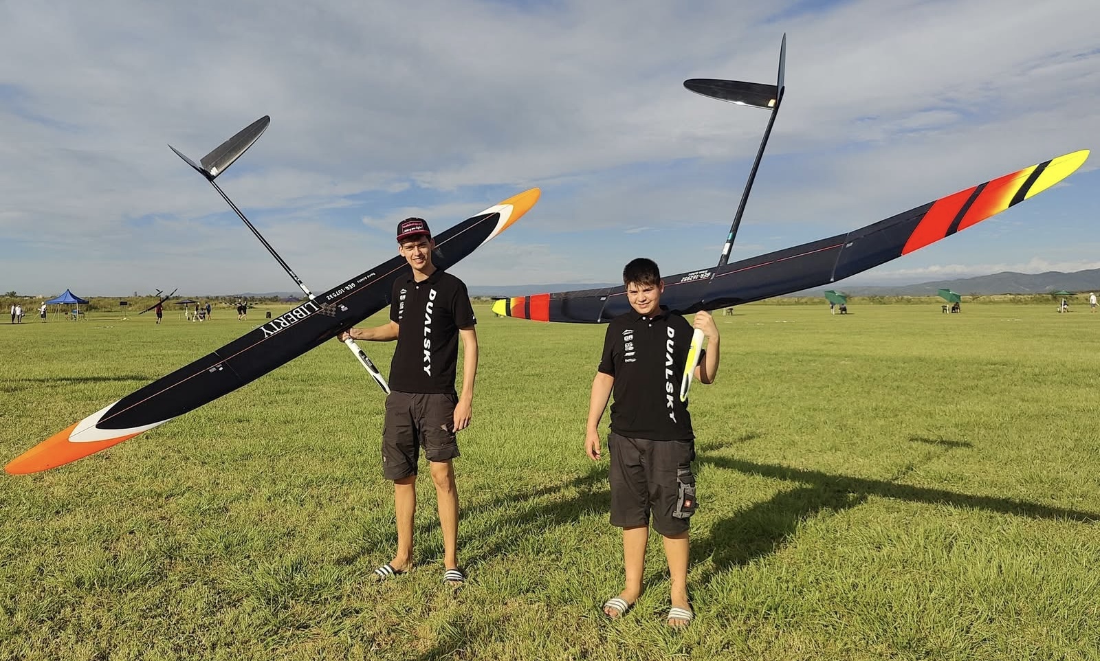 Zwei Modellflieger mit großen Segelflugmodellen auf Wiese, schwarze Shirts, bunte Flugzeuge mit langen Flügeln