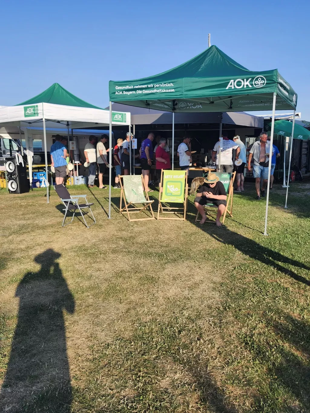 Grüne Pavillons mit Liegestühlen, dahinter warten Gäste an der Essensausgabe