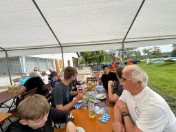 Mehrere Personen sitzen an Biertischen unter einem weißen Zelt, Bier und Getränke, Modellflugzeug im Hintergrund