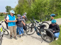 Fünf Radfahrer mit E-Bikes machen Pause auf einem Schotterweg, umgeben von Bäumen und Grünfläche