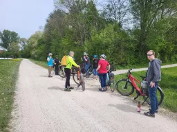 Mehrere Radfahrer mit Fahrrädern auf einem Schotterweg, umgeben von grüner Frühlingslandschaft mit Bäumen und Wiesen