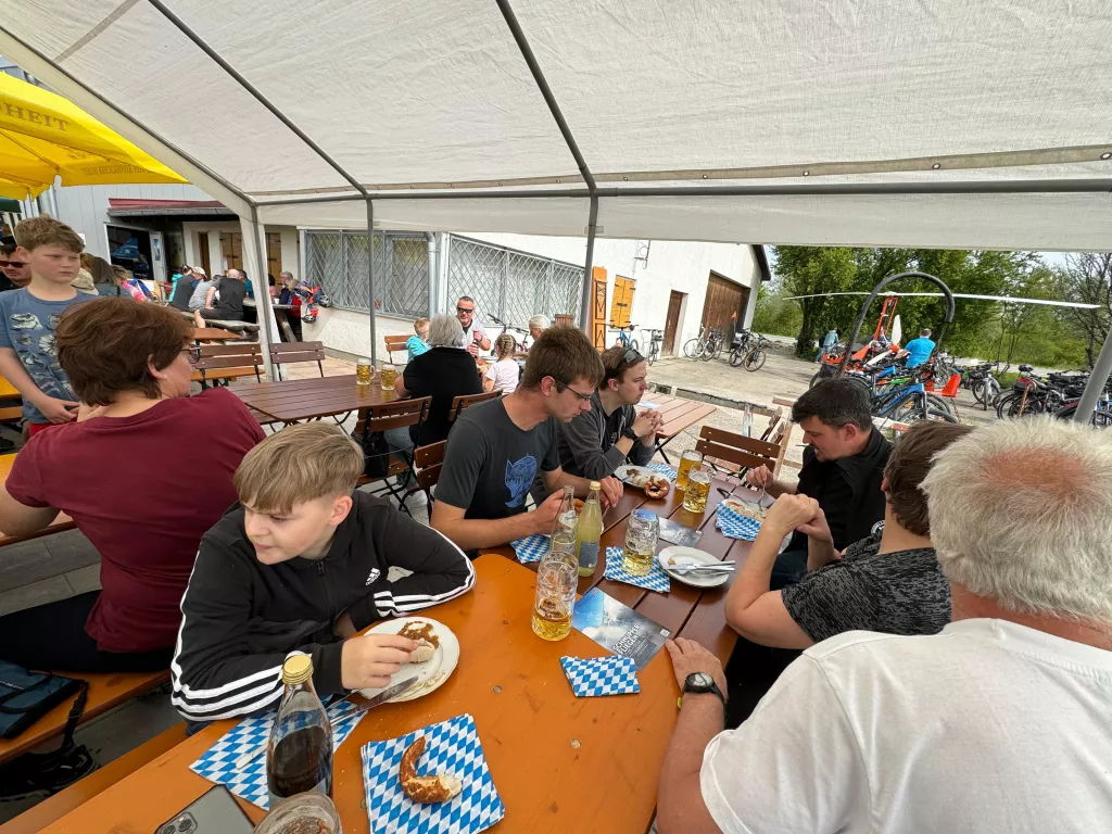 Überdachte Sitzgelegenheiten mit Besuchern, Brezeln und Bier auf blau-weißen Unterlagen, Fahrräder im Hintergrund