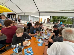 Überdachte Sitzgelegenheiten mit Besuchern, Brezeln und Bier auf blau-weißen Unterlagen, Fahrräder im Hintergrund