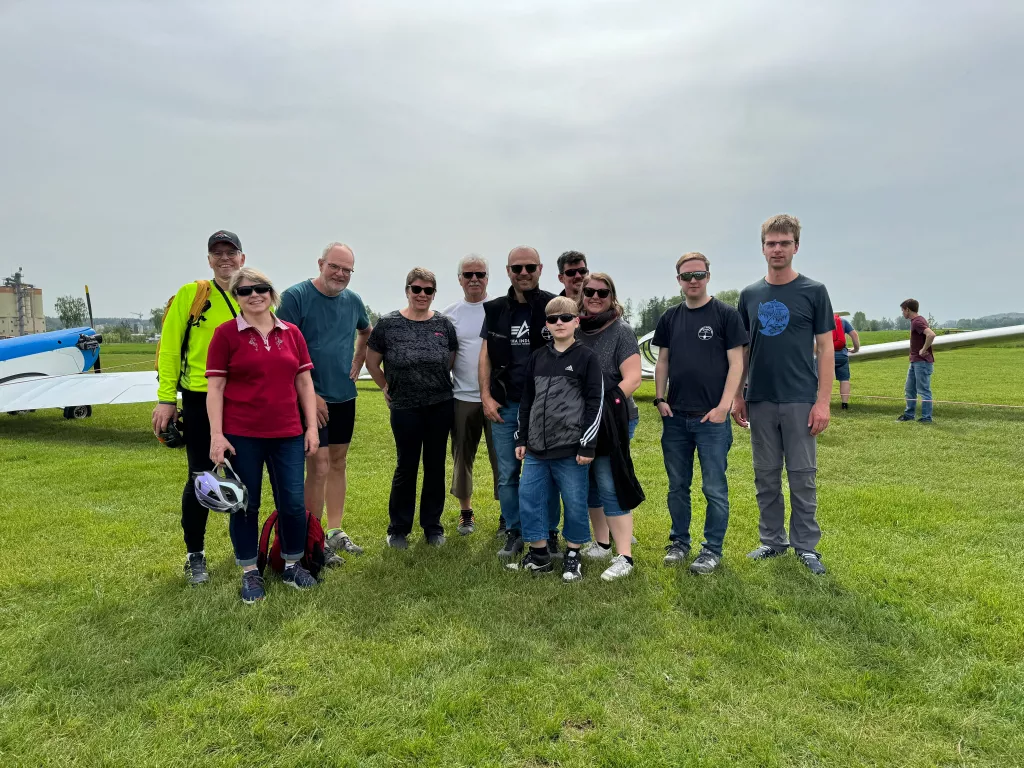 Zwölf Personen unterschiedlichen Alters stehen auf einer Wiese neben einem Flugzeug, bewölkter Himmel im Hintergrund