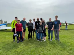 Zwölf Personen unterschiedlichen Alters stehen auf einer Wiese neben einem Flugzeug, bewölkter Himmel im Hintergrund