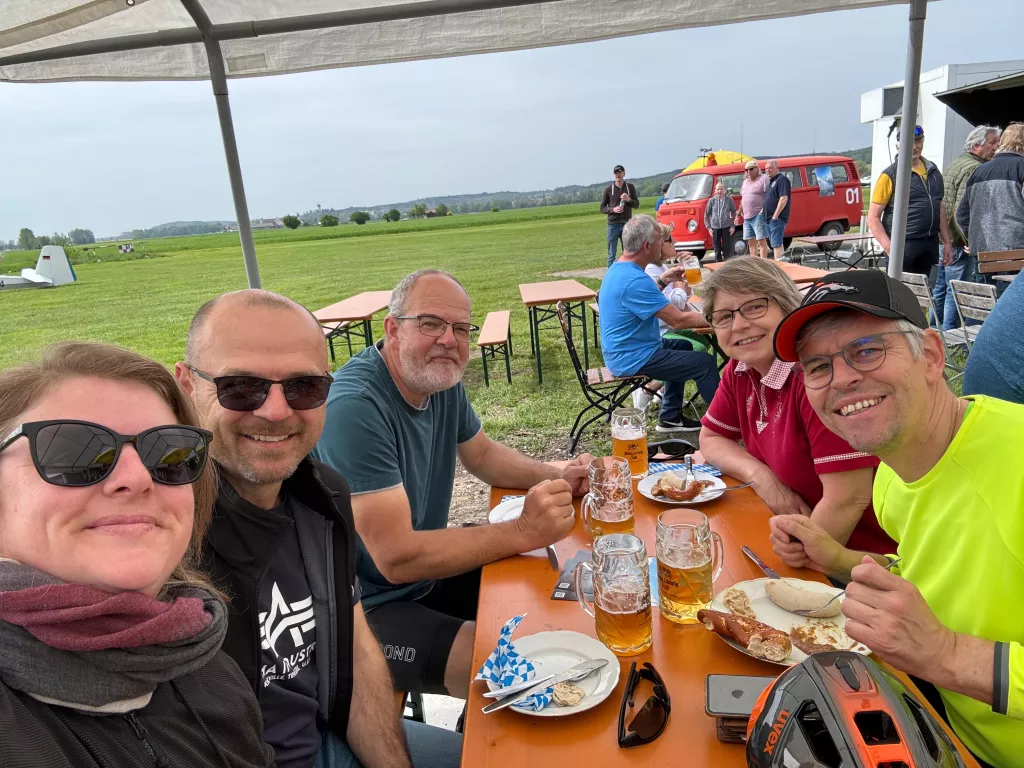 Fünf Personen an orangefarbener Bierbank, Biergläser, Flugplatz im Hintergrund, rotes Feuerwehrauto