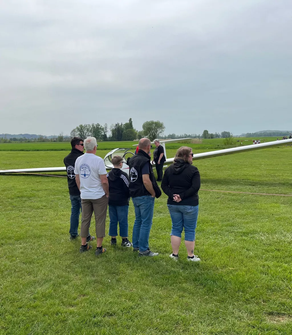 Gruppe von Personen steht auf Rasenfläche neben einem weißen Segelflugzeug, Landschaft im Hintergrund