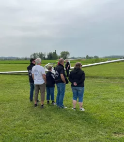 Gruppe von Personen steht auf Rasenfläche neben einem weißen Segelflugzeug, Landschaft im Hintergrund