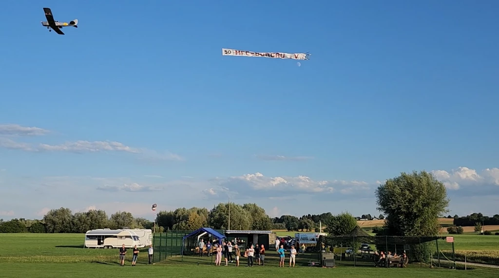 Modellflugzeug &uuml;ber gr&uuml;nem Rasenplatz, Vereinsgel&auml;nde mit Wohnwagen, Pavillon und Besuchern beim Sommerfest