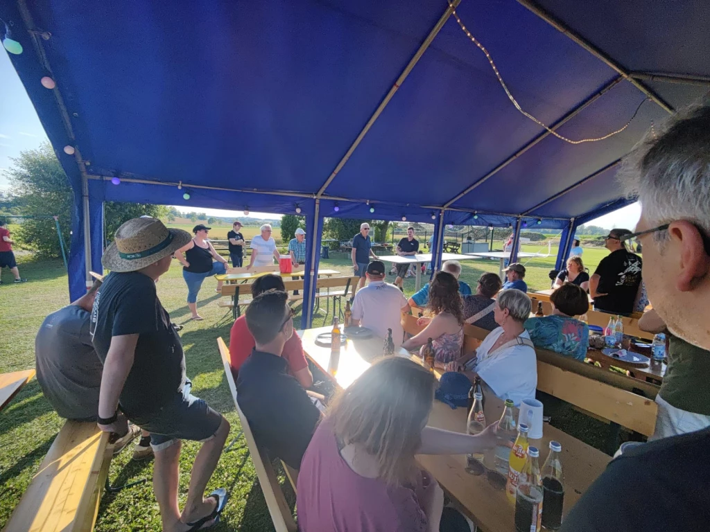 Gruppe von Menschen sitzt und steht unter einem blauen Partyzelt auf einer Wiese, Biertische, Vereinsveranstaltung