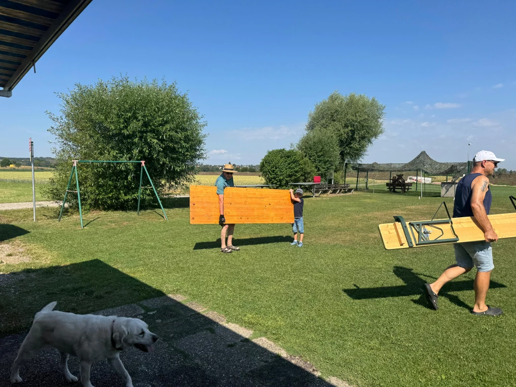 Zwei Personen tragen gelbes Holzbrett auf Rasenfl&auml;che, Hund im Schatten, Spielplatzschaukel im Hintergrund, sonniger Tag