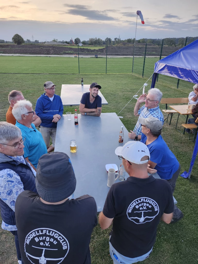 Mehrere M&auml;nner des Modellflugclubs Burgau sitzen an einem Tisch im Freien, Vereinsfahne und Zelt im Hintergrund