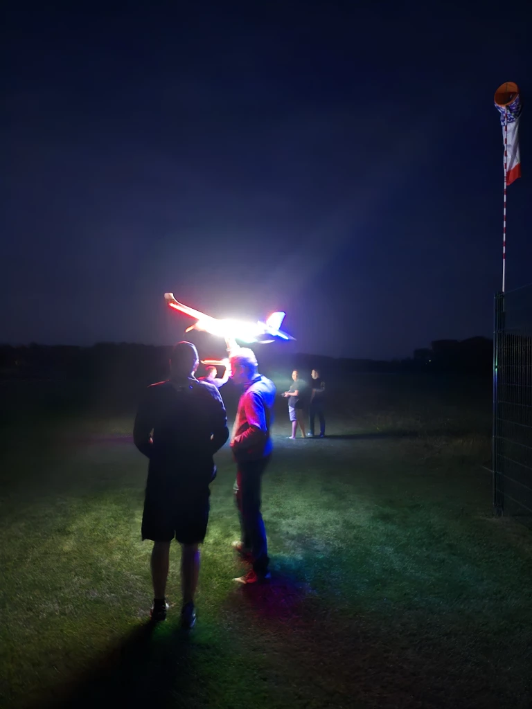 Abendszene auf Vereinsgelände: Zwei Personen beobachten beleuchtetes Modellflugzeug in Nachtkulisse mit Flagge