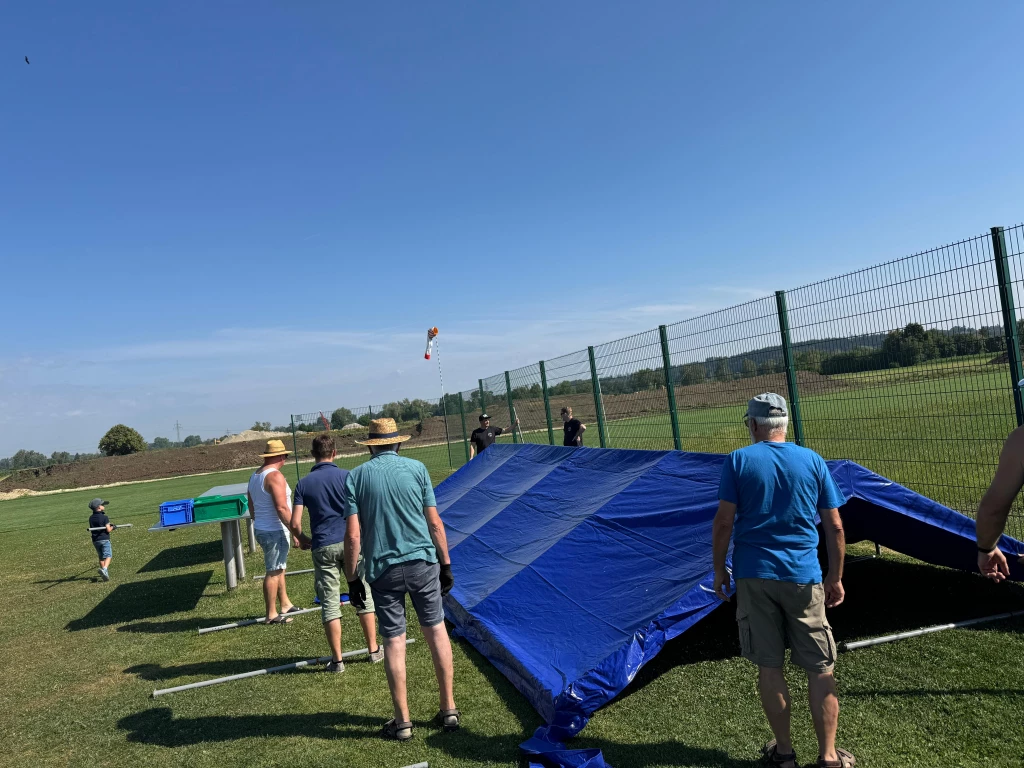 Mehrere Personen bewegen gro&szlig;e blaue Plane auf Modellflugplatz, Zaun und Wiese im Hintergrund