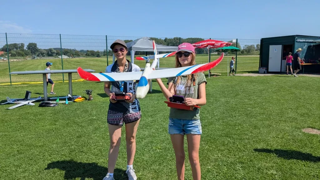 Zwei junge Frauen mit Modellflugsportbekleidung halten ein rot-blau-weißes Flugzeugmodell auf Rasenplatz mit Zelten