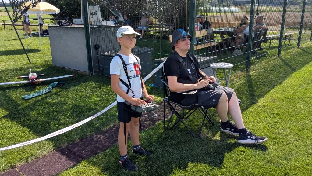 Zwei Personen am Modellflugplatz: Jugendlicher und Erwachsener mit Fernsteuerung, Modellflugzeug, grüne Rasenfläche