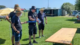Drei Männer in Vereinskleidung auf Rasenfläche, Holztisch im Vordergrund, gelber Sonnenschirm, Vereinscontainer im Hintergrund