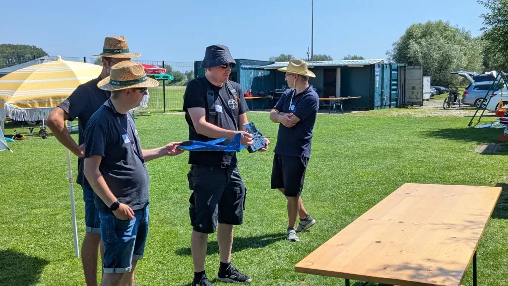 Drei Personen mit Sonnenhüten an einem Tisch auf Rasenplatz, Vereinscontainer im Hintergrund, Sommertag