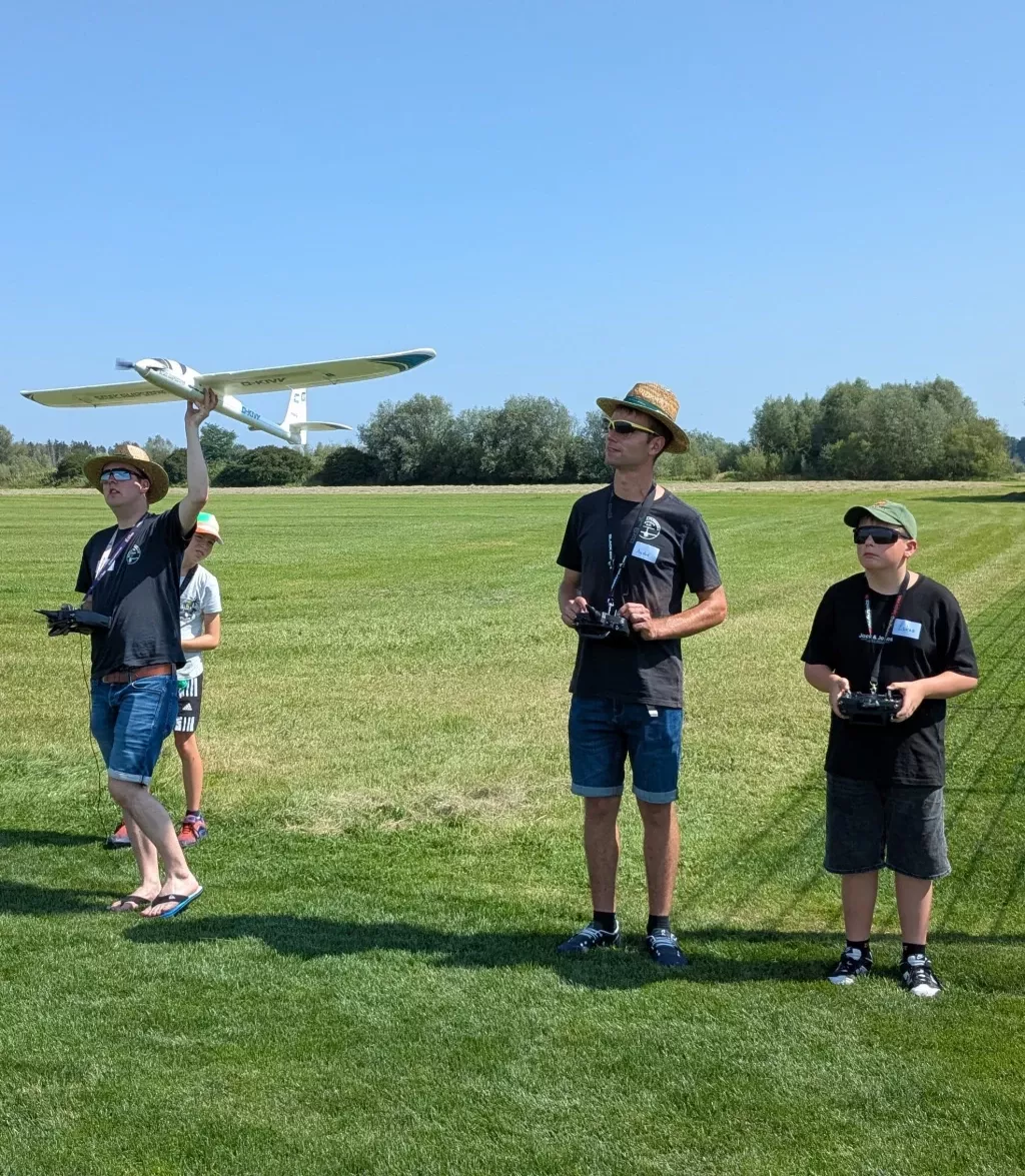 Vier Personen auf einer grünen Wiese mit Segelflugmodell, zwei halten Fernsteuerungen, einer präsentiert ein weißes Flugzeugmodell