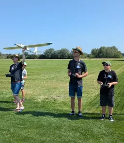 Vier Personen auf einer grünen Wiese mit Segelflugmodell, zwei halten Fernsteuerungen, einer präsentiert ein weißes Flugzeugmodell