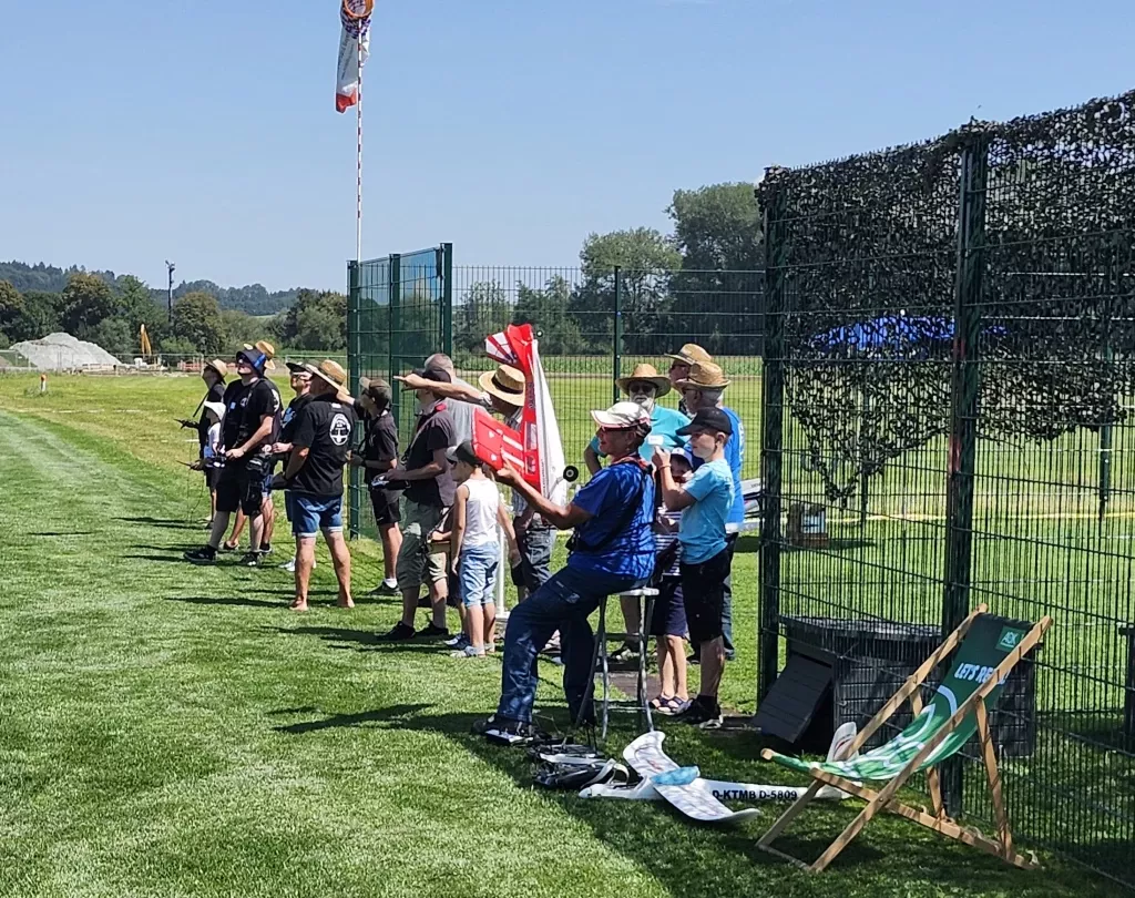 Gruppe von Menschen an einem sonnigen Tag auf einem Modellflugplatz, stehen entlang eines grünen Rasens, Zaun im Hintergrund