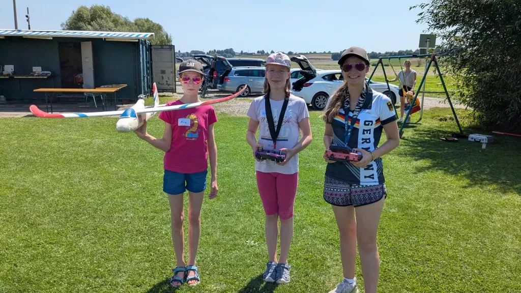Drei junge Teilnehmerinnen mit Modellflugzeugen und Fernsteuerungen auf Vereinsgelände, Sommertag