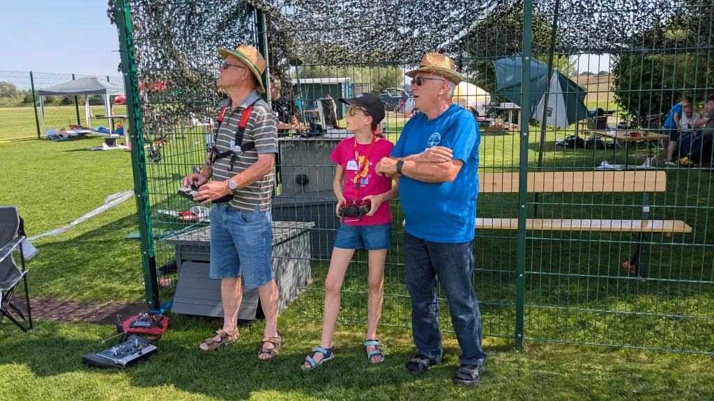 Drei Personen mit Fernsteuerungen beim Modellflug: Zwei Männer und ein junges Mädchen an einem Zaun auf Rasenplatz