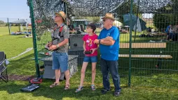 Drei Personen mit Fernsteuerungen beim Modellflug: Zwei Männer und ein junges Mädchen an einem Zaun auf Rasenplatz