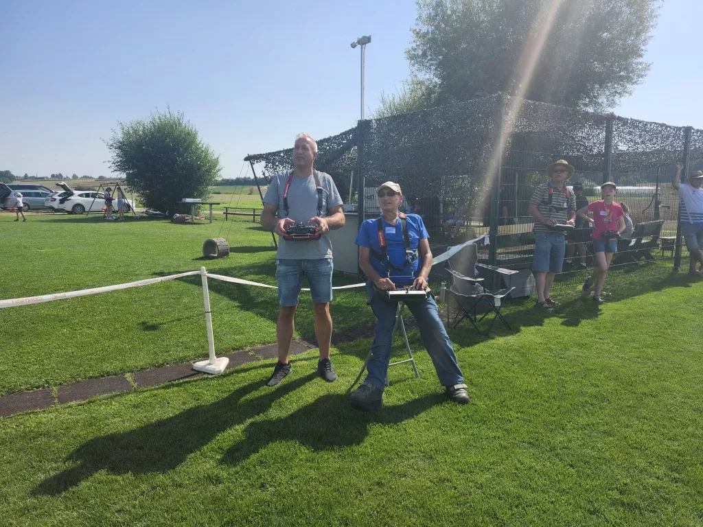 Zwei Modellflug-Enthusiasten mit Fernsteuerungen auf grünem Rasenplatz, Zaun und Bäume im Hintergrund