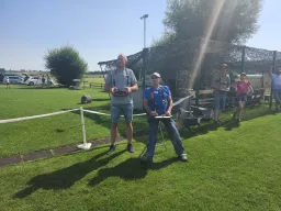 Zwei Modellflug-Enthusiasten mit Fernsteuerungen auf grünem Rasenplatz, Zaun und Bäume im Hintergrund