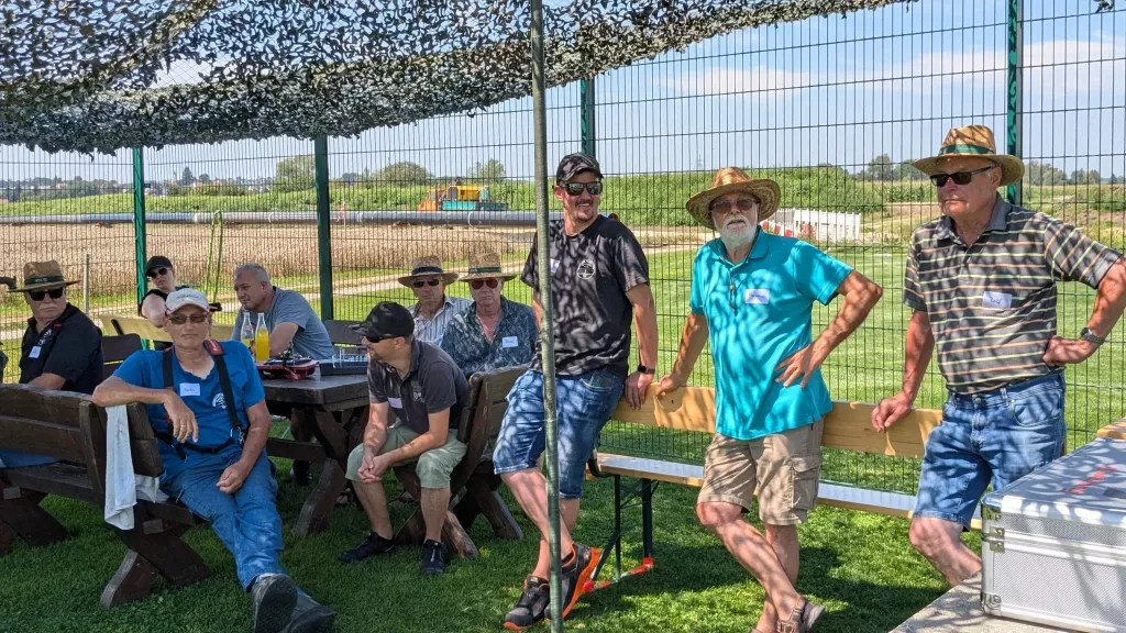 Gruppe von Männern in Sommerkleidung auf Rasenfläche mit Flugplatzkulisse, Sitzbänke und Sonnenschutz