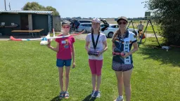 Drei junge Teilnehmerinnen mit Modellflugzeugen und Fernsteuerungen auf Vereinsgelände, Sommertag