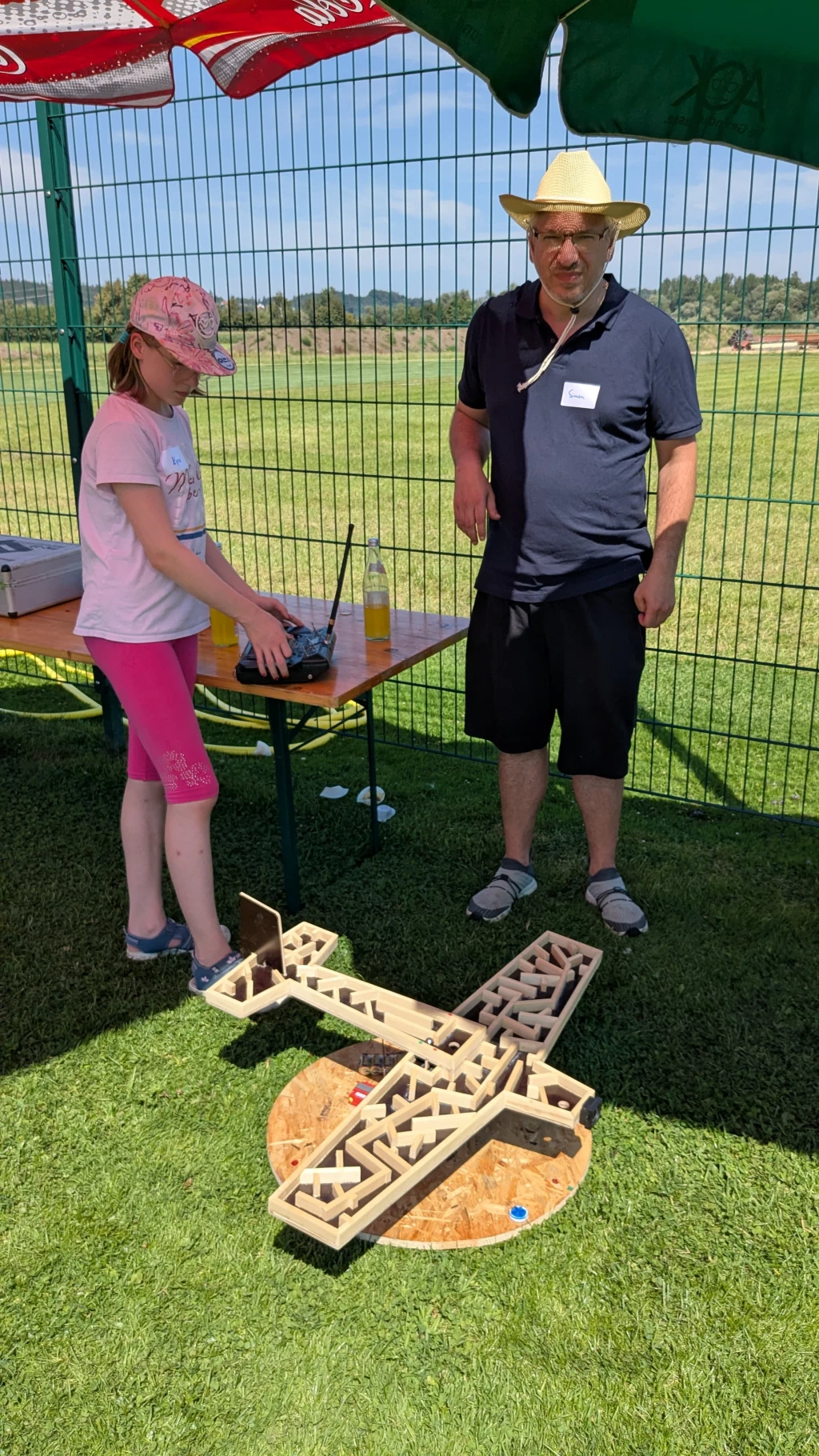 Mädchen und Mann mit Strohhut stehen an einem Tisch mit einem großen Holzflugzeug-Modell auf grünem Rasen, umgeben von Zaun