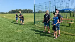 Fünf Personen mit Fernsteuerungen auf Modellflugplatz, grüne Wiese, Zaun, Bäume im Hintergrund